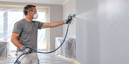 pro-painter-fine-finish Professional painter wearing a respirator applies an even wall coating with an airless spray gun